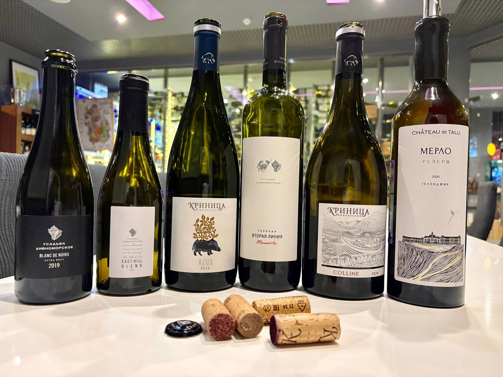 Несколько бутылок вина в ряд (Криница, Усадьба Дивноморское, Chateau de Talu) на столе и пробки на переднем плане.