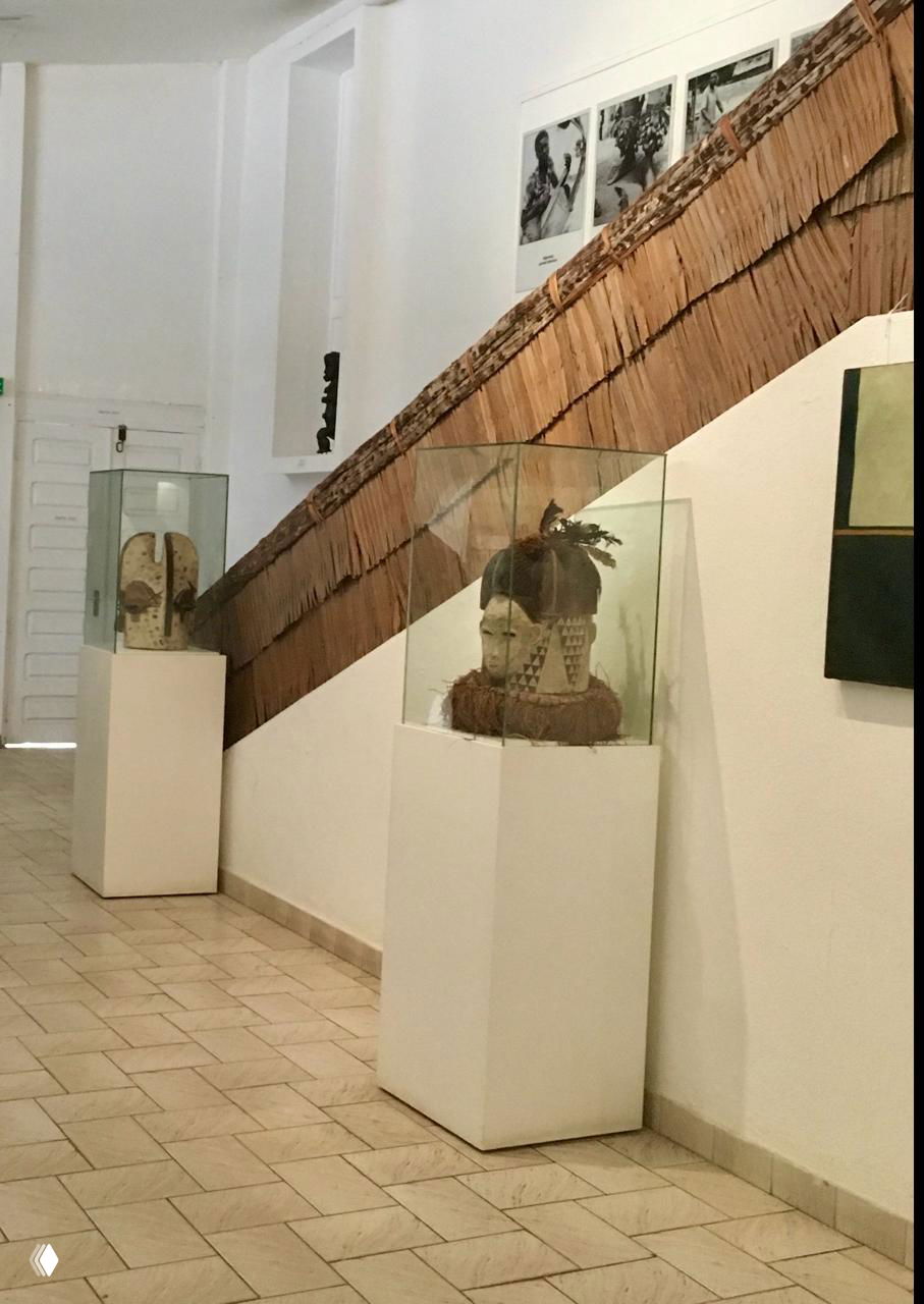 Витрина музея с двумя головными уборами и масками в стеклянных колпаках на белых постаментах; интерьер галереи, экспонаты под лампами.