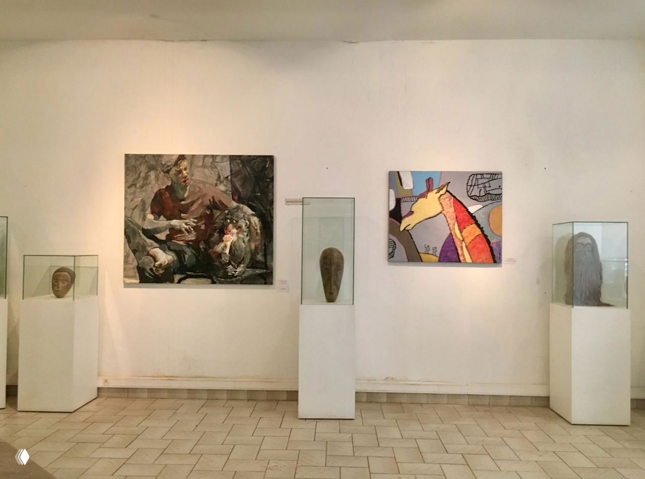 Интерьер выставочной залы: картины на стенах и несколько масок в стеклянных витринах на белых постаментах, организованная экспозиция галереи.