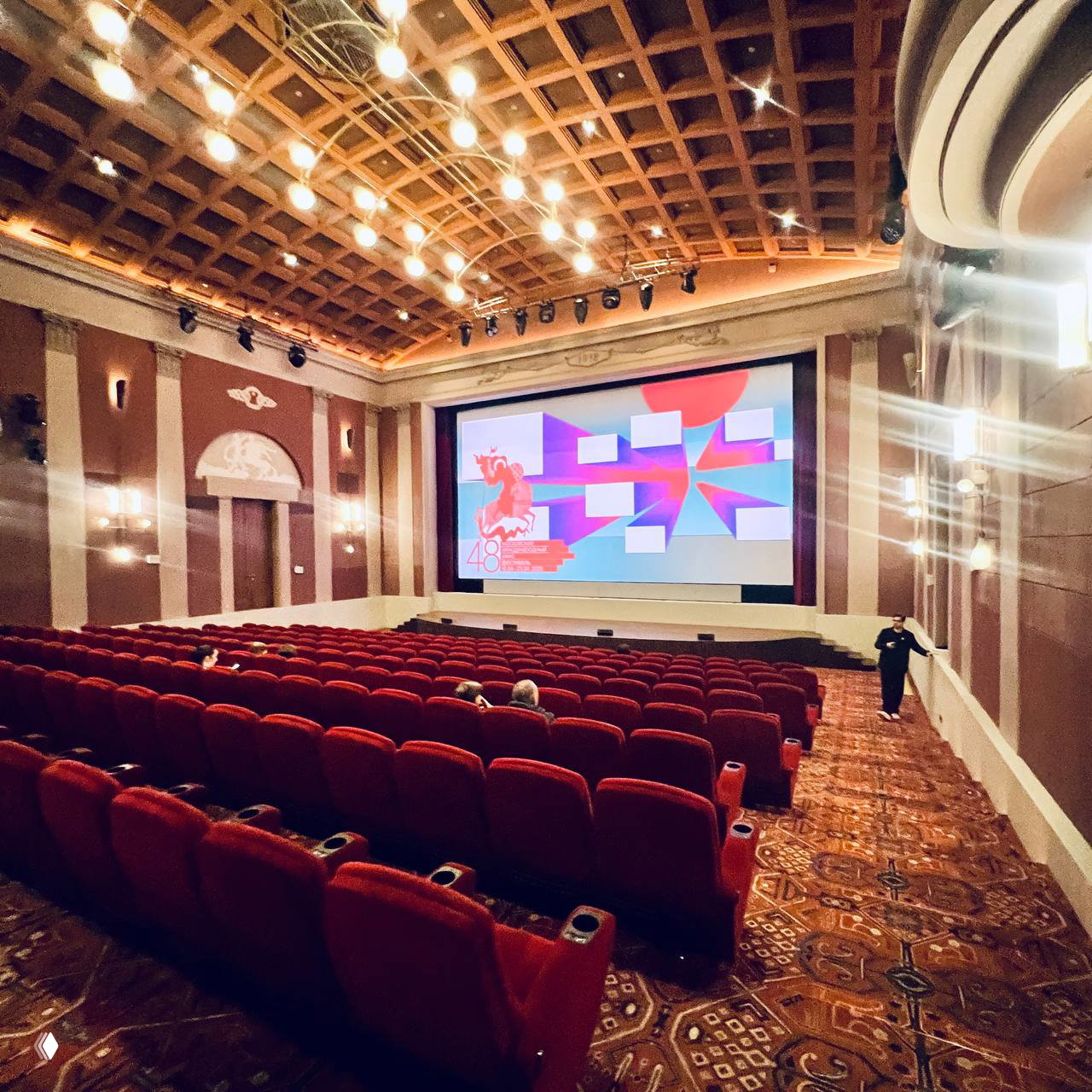 Пустой кинозал с красными креслами и большим экраном, на котором отображается графика и логотип 48-го Московского кинофестиваля; интерьер зрительного зала.