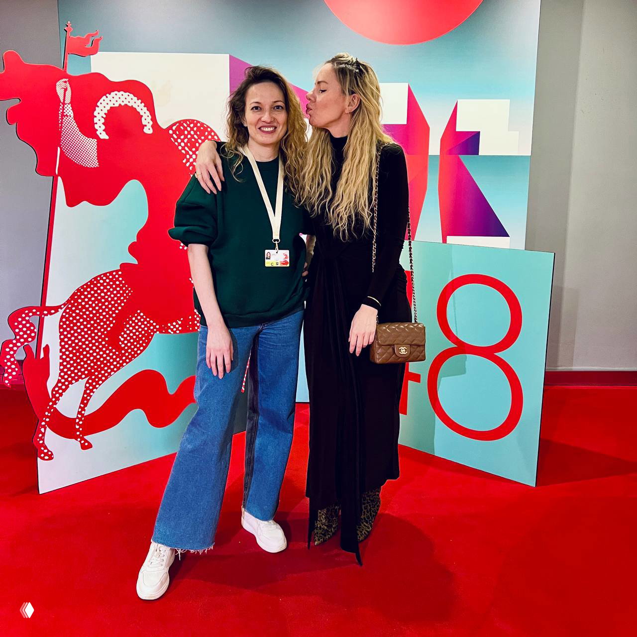 Две женщины позируют на красной дорожке перед пастельной графикой фестиваля с цифрой 48; одна дарит поцелуй в щеку другой — дружеская сцена на фоне инсталляции.