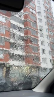 Кадр из салона автомобиля через дождливое боковое стекло: крупные капли искажают фасад многоэтажного дома и городскую улицу.