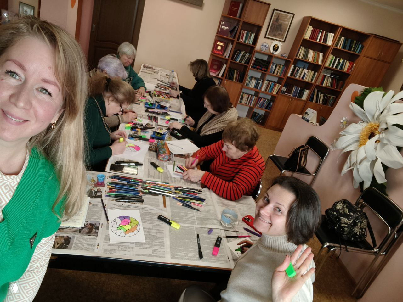 Селфи ведущей в зелёной жилетке на фоне длинного стола, где участницы раскрашивают нейрографику; видны фломастеры, карандаши и листы с эскизами.