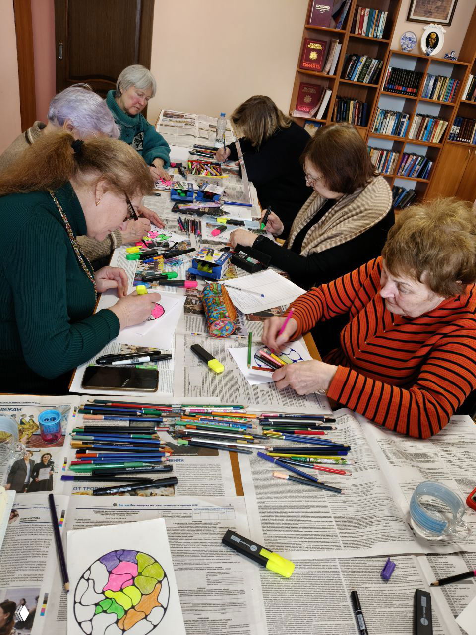 Участницы за столом сосредоточенно раскрашивают нейрографические эскизы; на столе много карандашей, маркеров и газет, кадр снят сбоку.