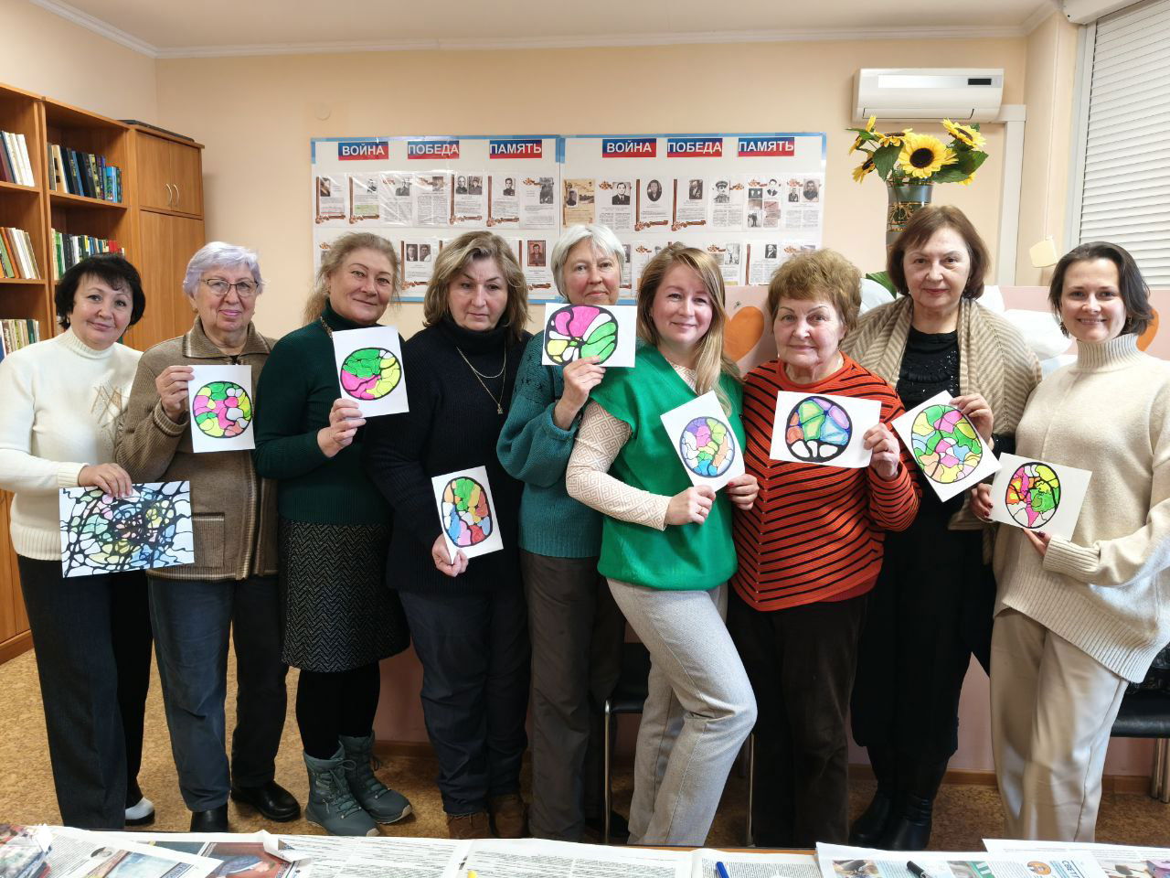 Групповая фотография женщин пожилого возраста в Центре общения Светлогорска: они держат свои яркие рисунки нейрографики и улыбаются ведущей.