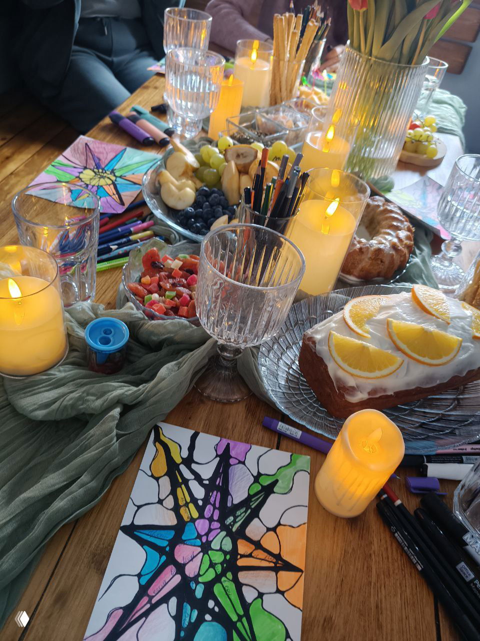 Фрагмент стола с готовой работой нейрографики, подсвечниками и стаканами; видны маркеры, фрукты и десерты.