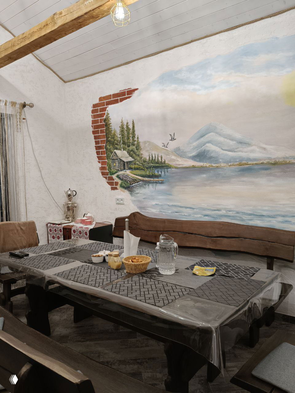 Комната отдыха с настенной фреской: стол застелен, на столе закуски и графин воды, деревянные лавки и большая пейзажная роспись.