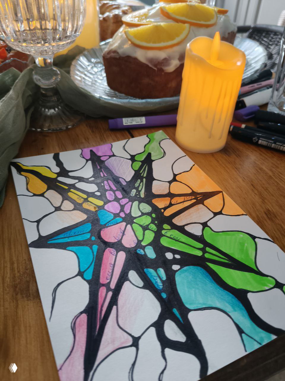 Лист с нейрографическим рисунком в виде звезды, раскрашенным маркерами; на столе свеча, кусочек торта, стеклянная посуда и художественные материалы.