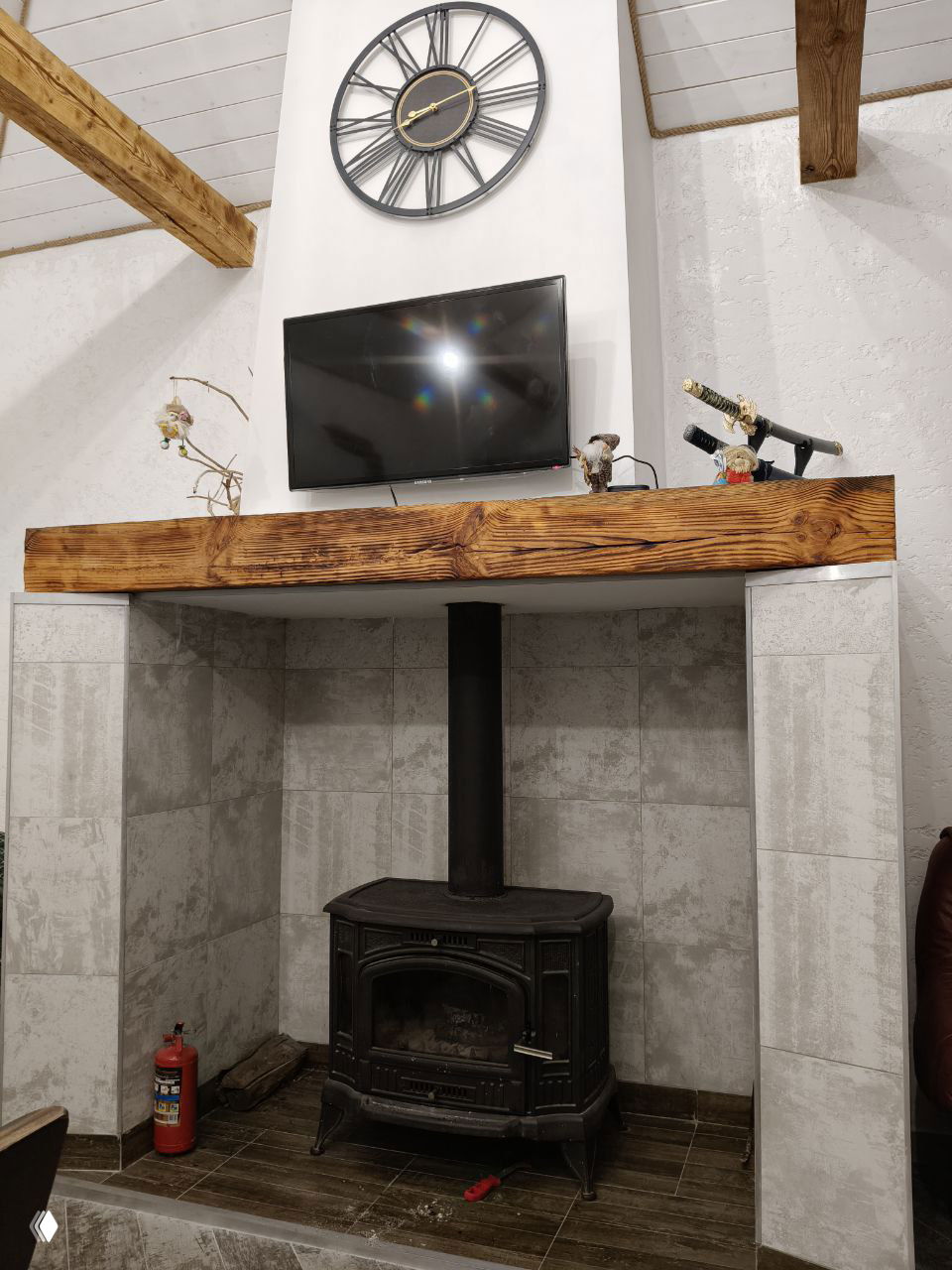 Дровяная чугунная печь в предбаннике: печь с дымоходом, над ней телевизор на стене и крупные деревянные балки в интерьере.