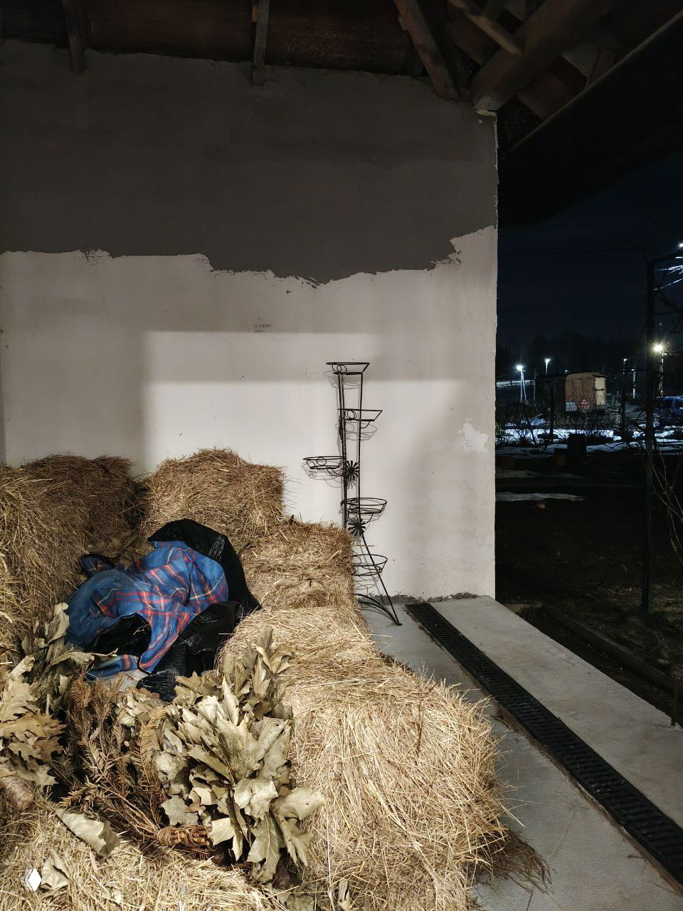 Склад тюков сена у бани: тюки сена, рядом пучки веников и рабочая одежда; вид на открытое помещение и ночной двор с освещением.