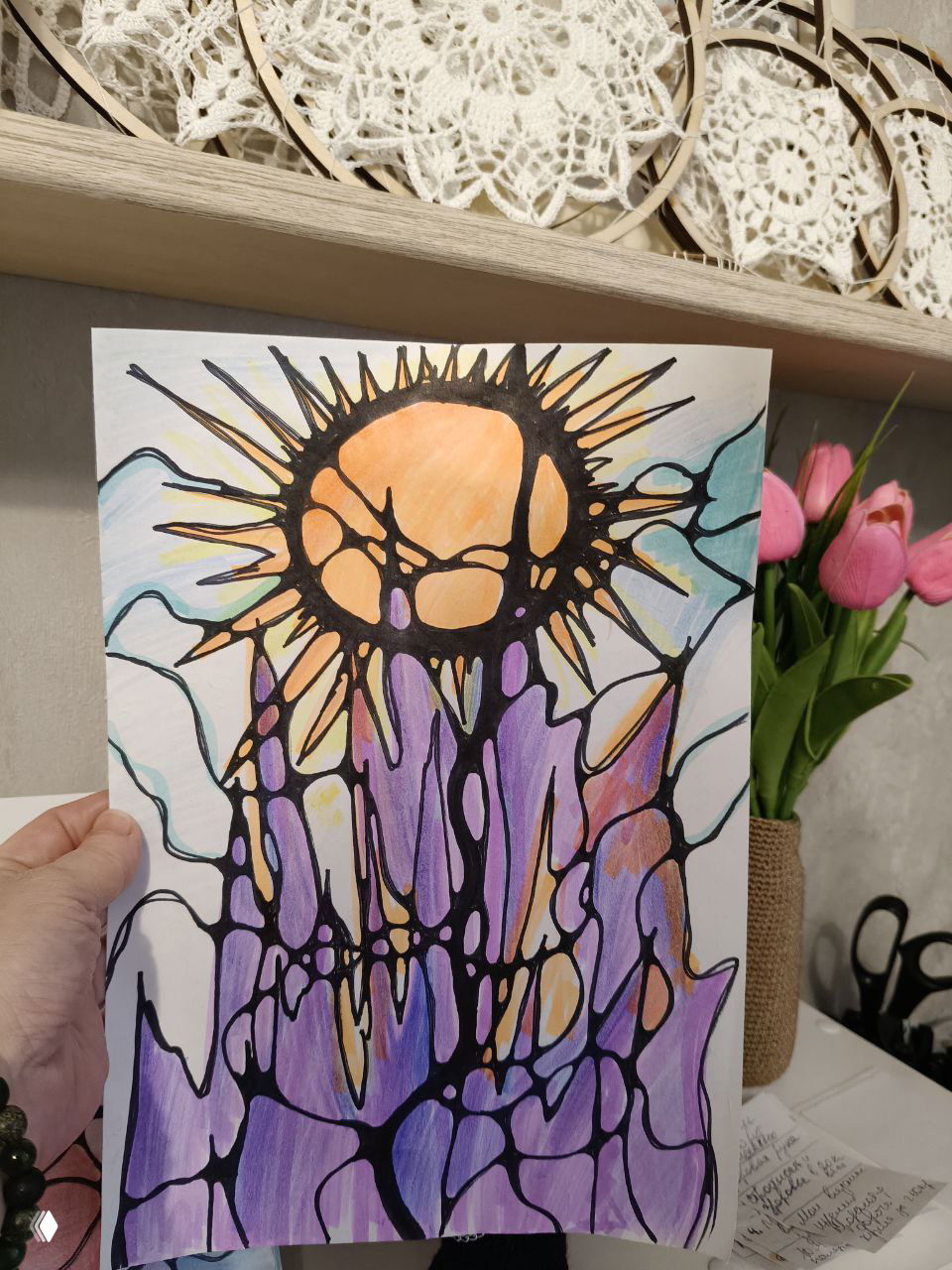 Рука показывает нейрографическую работу с солнечным мотивом и вытянутыми линиями; за работой видны тюльпаны и декоративные салфетки на полке.