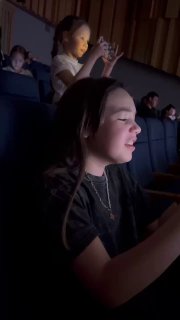 Крупный план племянницы в тёмном зале: девушка в профиль, подсвеченная сценой, поёт и улыбается, эмоциональный момент во время концерта.