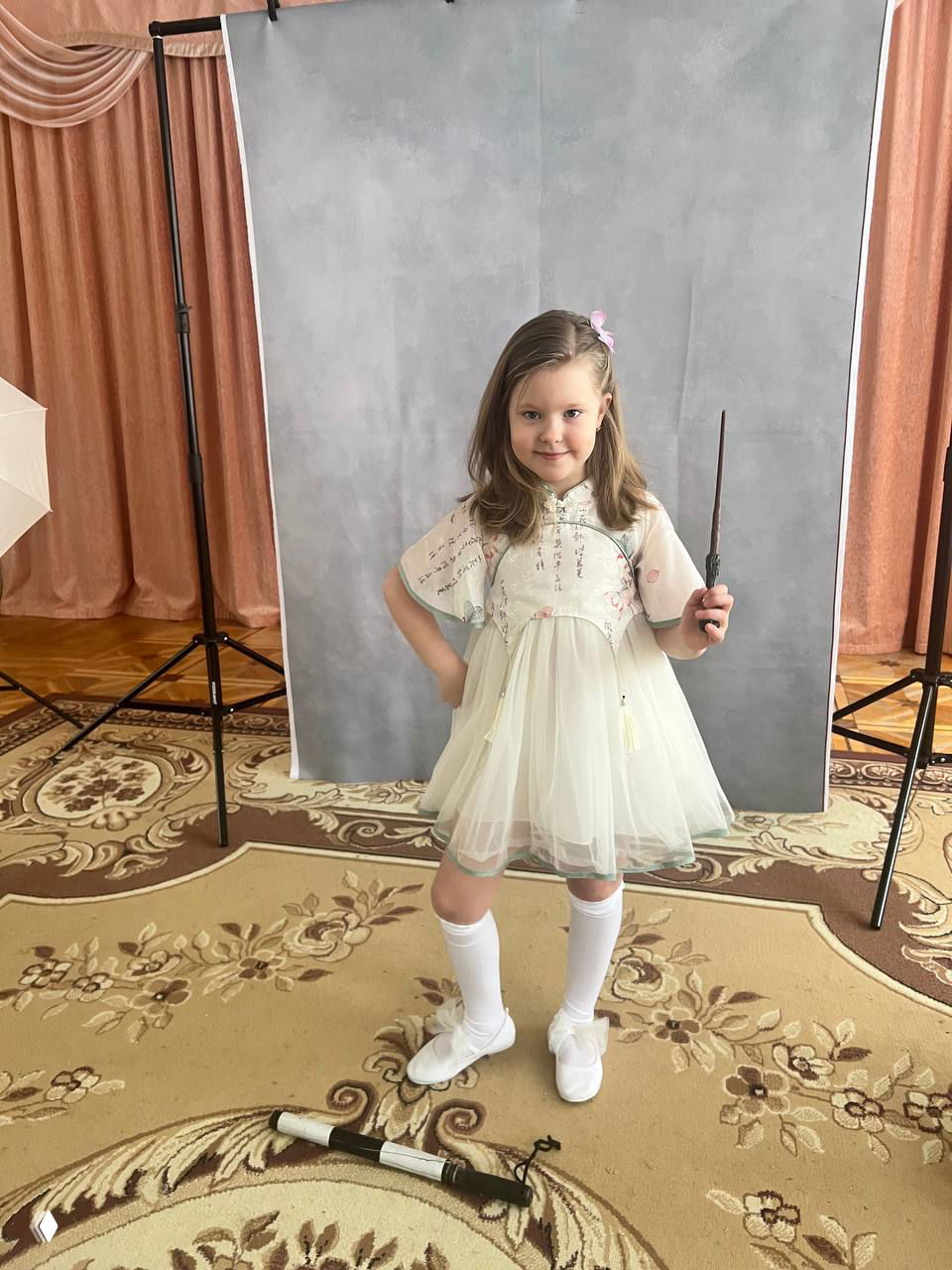 Девочка в белом платье и гольфах позирует на фоне студийного фона в детском саду во время фотосессии — детская улыбка и наряд для выпускного.