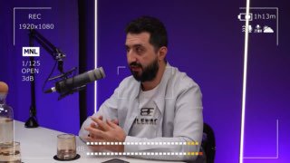 Человек за столом в подкаст-студии на фиолетовом фоне с микрофоном, камерой и визуальными элементами интерфейса записи в кадре