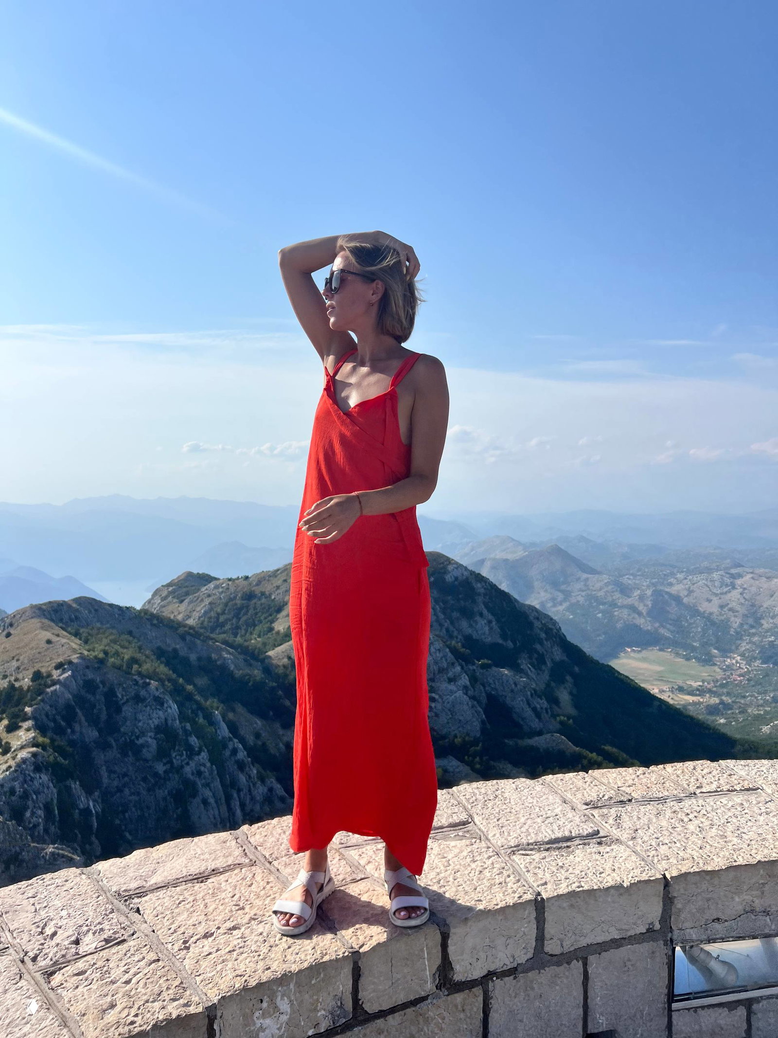 Фигура женщины в красном платье на каменной ограде вершины, фон — далеко уходящие горы и прозрачное голубое небо, ощущение простора.