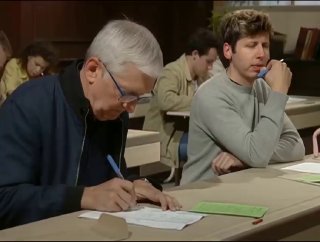 Two men seated at desks in a classroom-like setting, concentrating on writing on paper; older man foreground, others blurred in the background.