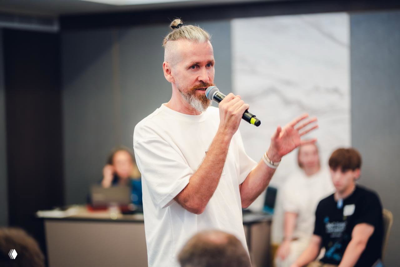 Speaker Sergey Osipov on stage speaking into a microphone at the hackathon, addressing participants in a conference room setting with focus.