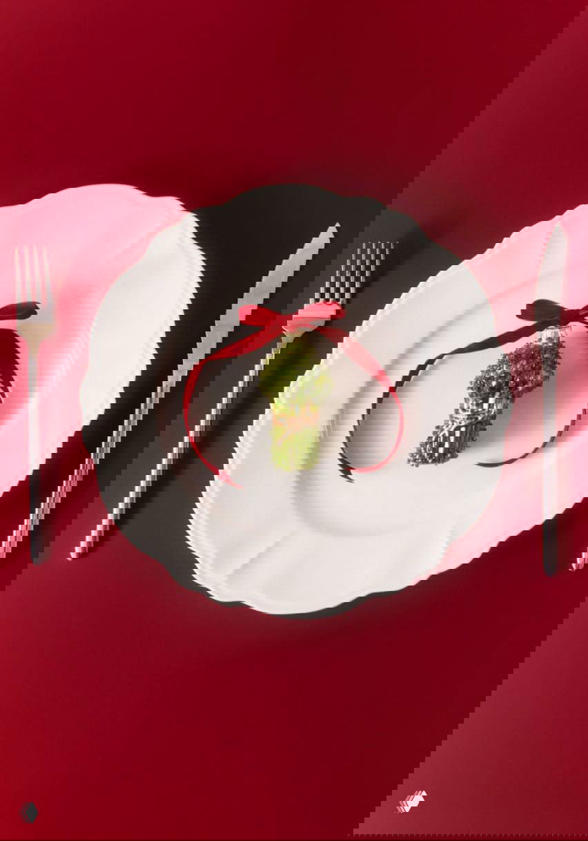 Ёлочная игрушка-брокколи из стекла, украшена красной ленточкой, лежит на белой тарелке с прибором на бордовом фоне.