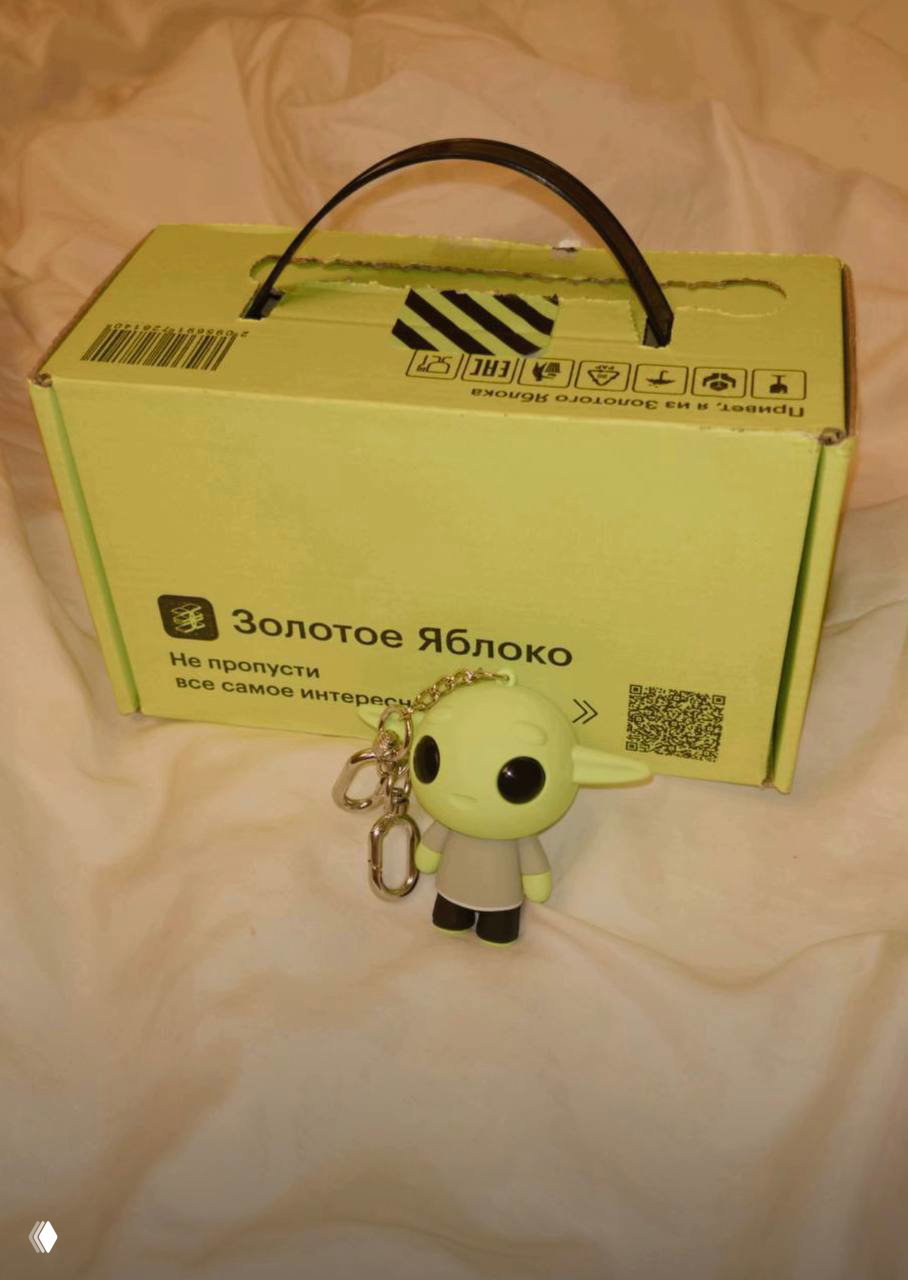 Салатовая коробка «Золотое Яблоко» на постели, перед ней брелок в виде зеленого эльфа — распаковка и содержимое.