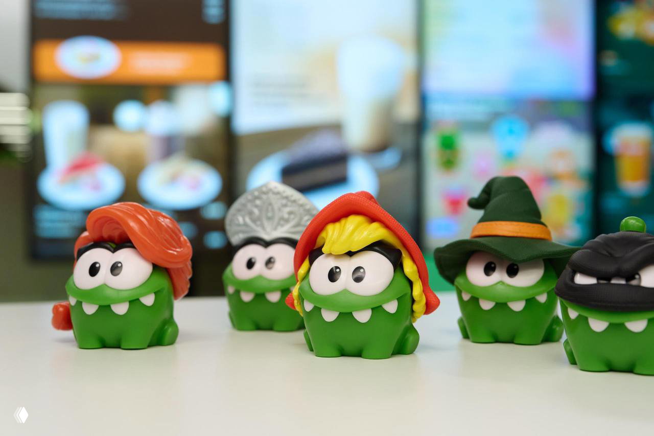 Группа маленьких зелёных фигурок Ам Нямов в образах Красной шапочки, Снегурочки, ведьмы и других персонажей на фоне меню кафе.
