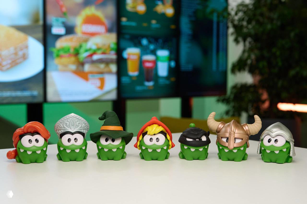 Ряд коллекционных фигурок Ам Нямов в различных костюмах — богатырь, ниндзя, викинг, волшебник и женские версии, выстроенные на столе в кафе.