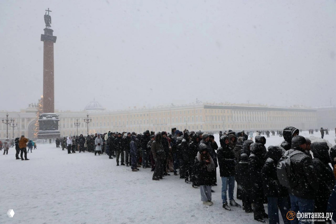 Крупный план плотной очереди у Эрмитажа: люди в зимних куртках и шапках стоят в линию на заснеженной площади рядом с Александровской колонной.