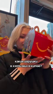 Человек в ресторане McDonald's держит коробку Хэппи Мил с оформлением «Зверополис 2», видна часть интерьера и окно с улицей за ним.