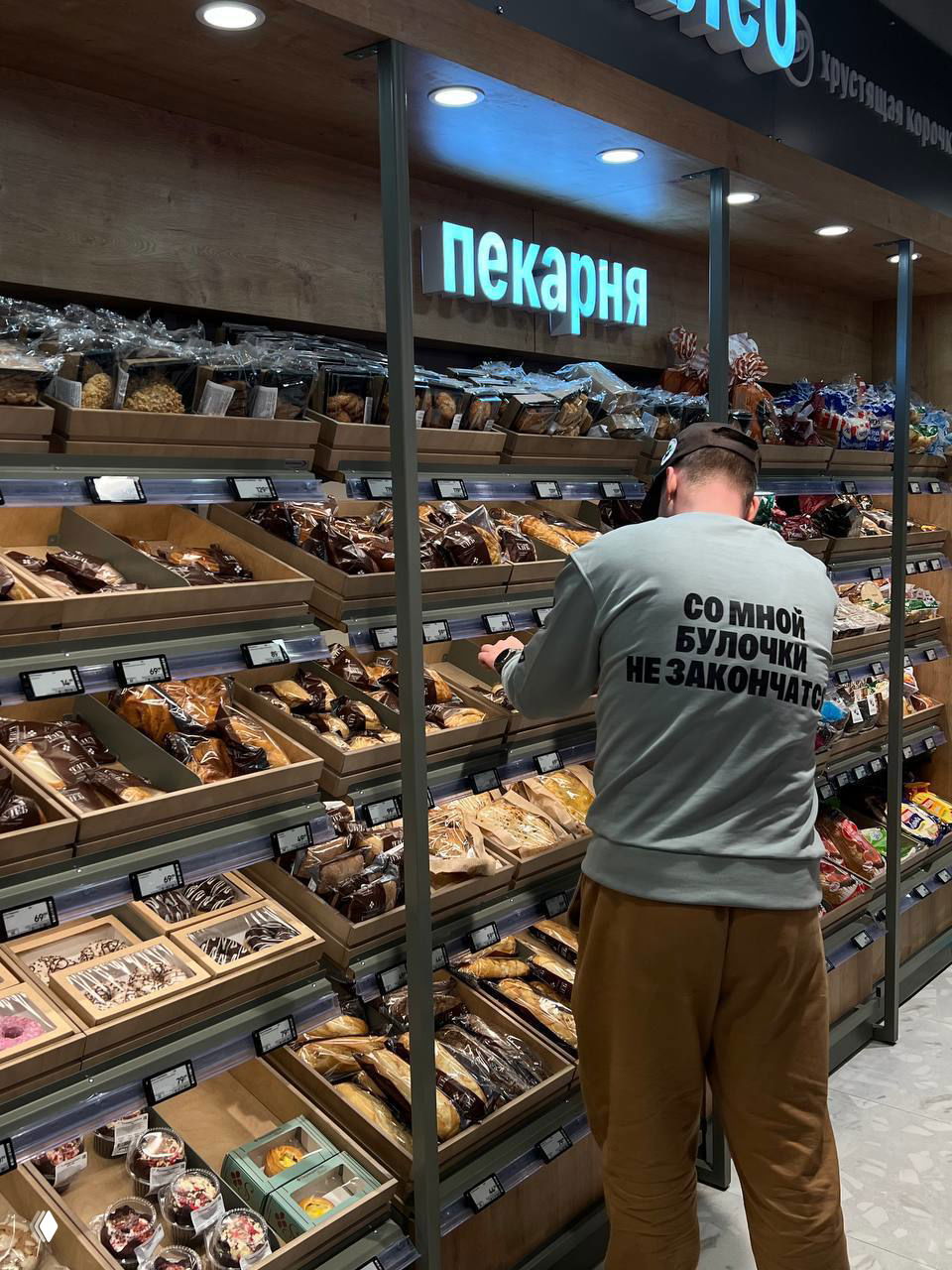 Отдел пекарни в Select: сотрудник спиной раскладывает выпечку на полках, на толстовке крупная юмористическая надпись про булочки, видны полки с хлебобулочными изделиями.