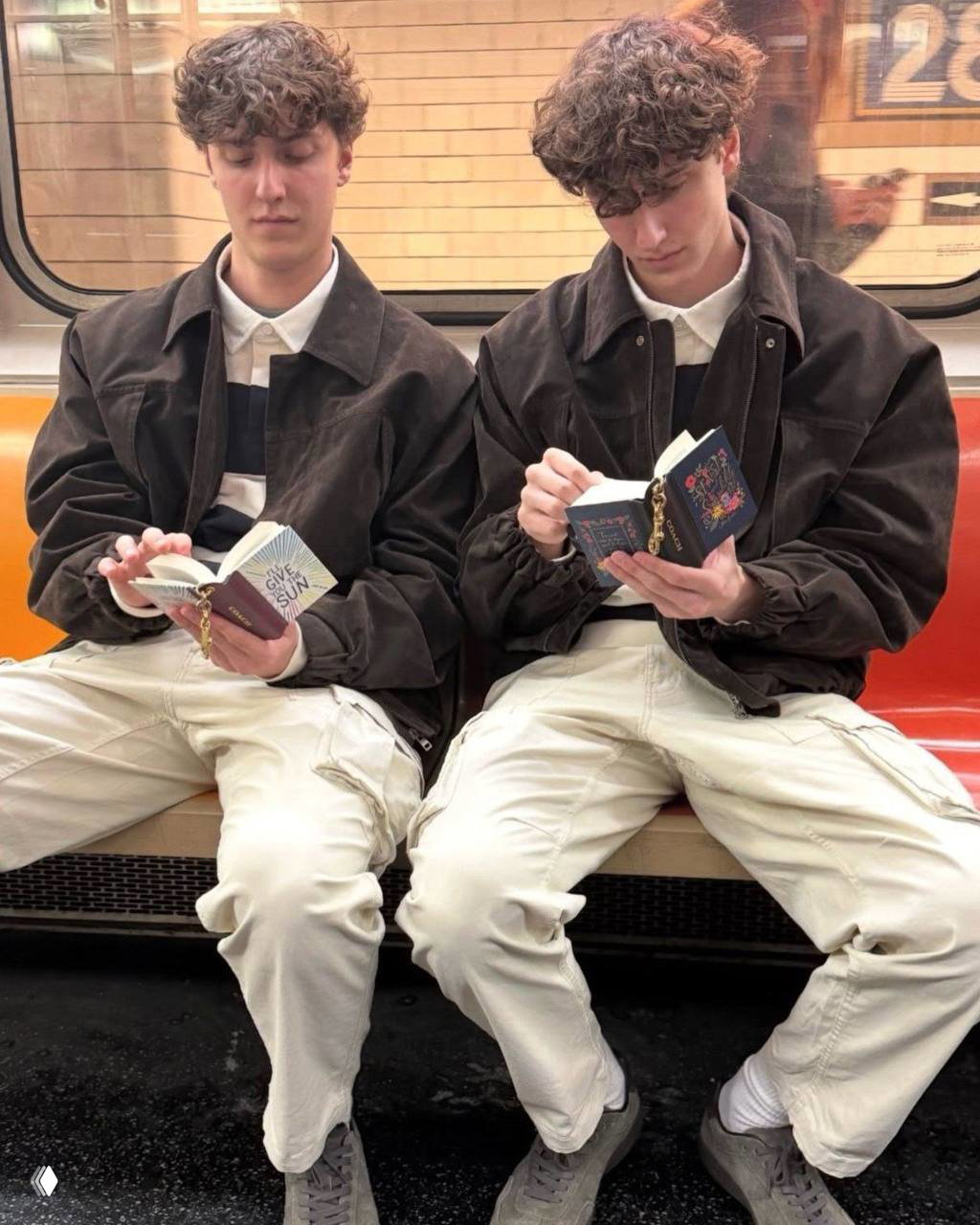 Два молодых человека в метро читают мини‑книжки‑подвески Coach — маленькие книги на цепочках, прикреплённые к сумкам, повседневная городская сцена.