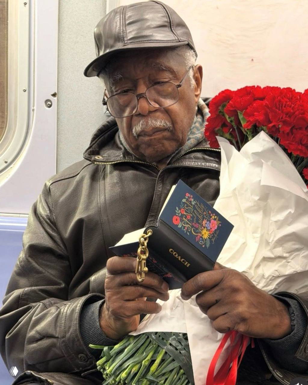 Пожилой пассажир в метро внимательно читает мини‑книжку Coach, держит букет цветов; кадр подчёркивает универсальность и доступность аксессуара для чтения.