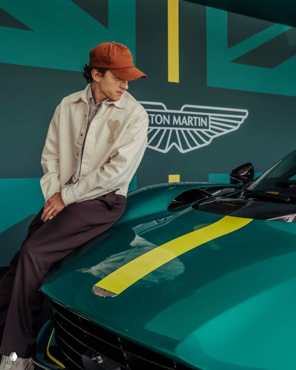 Том Холланд опирается на капот зелёного Aston Martin с жёлтой полосой; на заднем плане логотип Aston Martin — кадр из кампейна.
