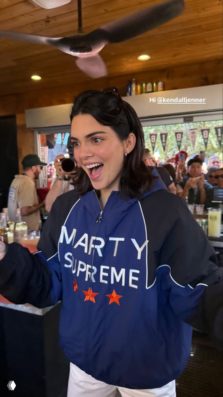 Женщина улыбается в баре, одета в синюю брендированную куртку с текстом на груди; внутри видна толпа и интерьер заведения.