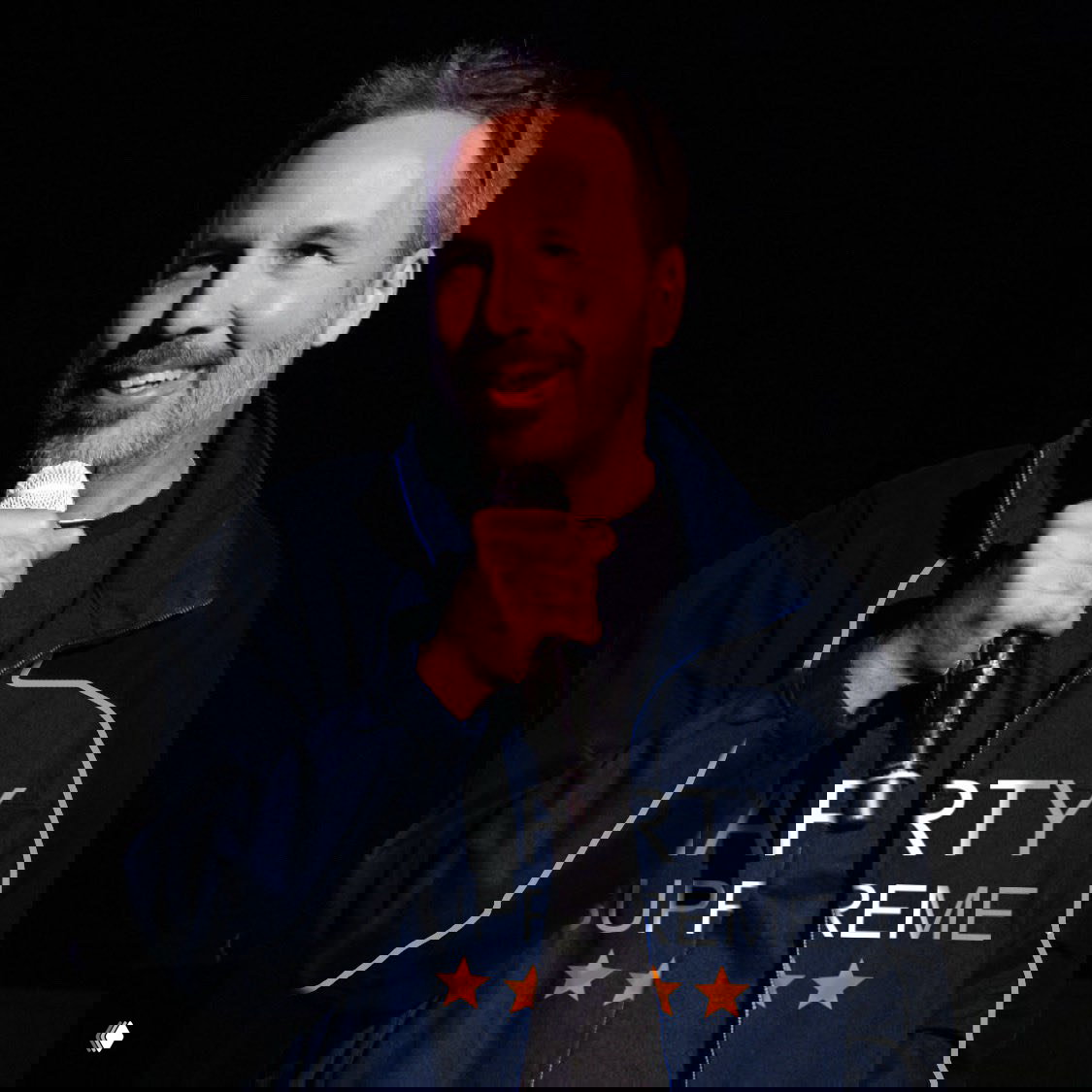 Человек на сцене держит микрофон, на нём тёмно-синяя куртка с крупной надписью и звёздами — кадр с выступления в тёмном зале.