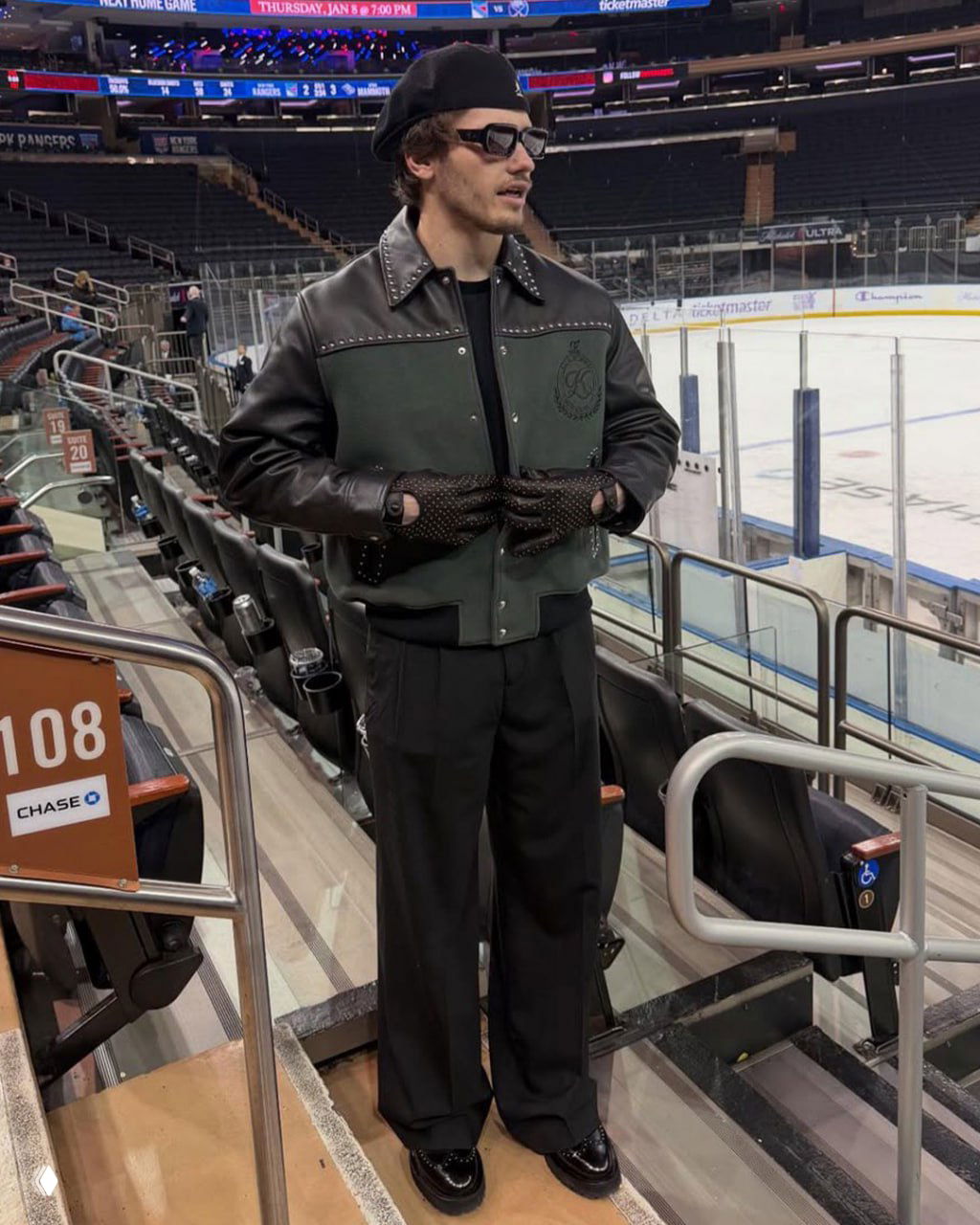 Мужчина в двухцветной кожаной куртке со заклёпками и перчатках стоит на трибуне ледовой арены, виден лёд и ряд кресел вокруг него.