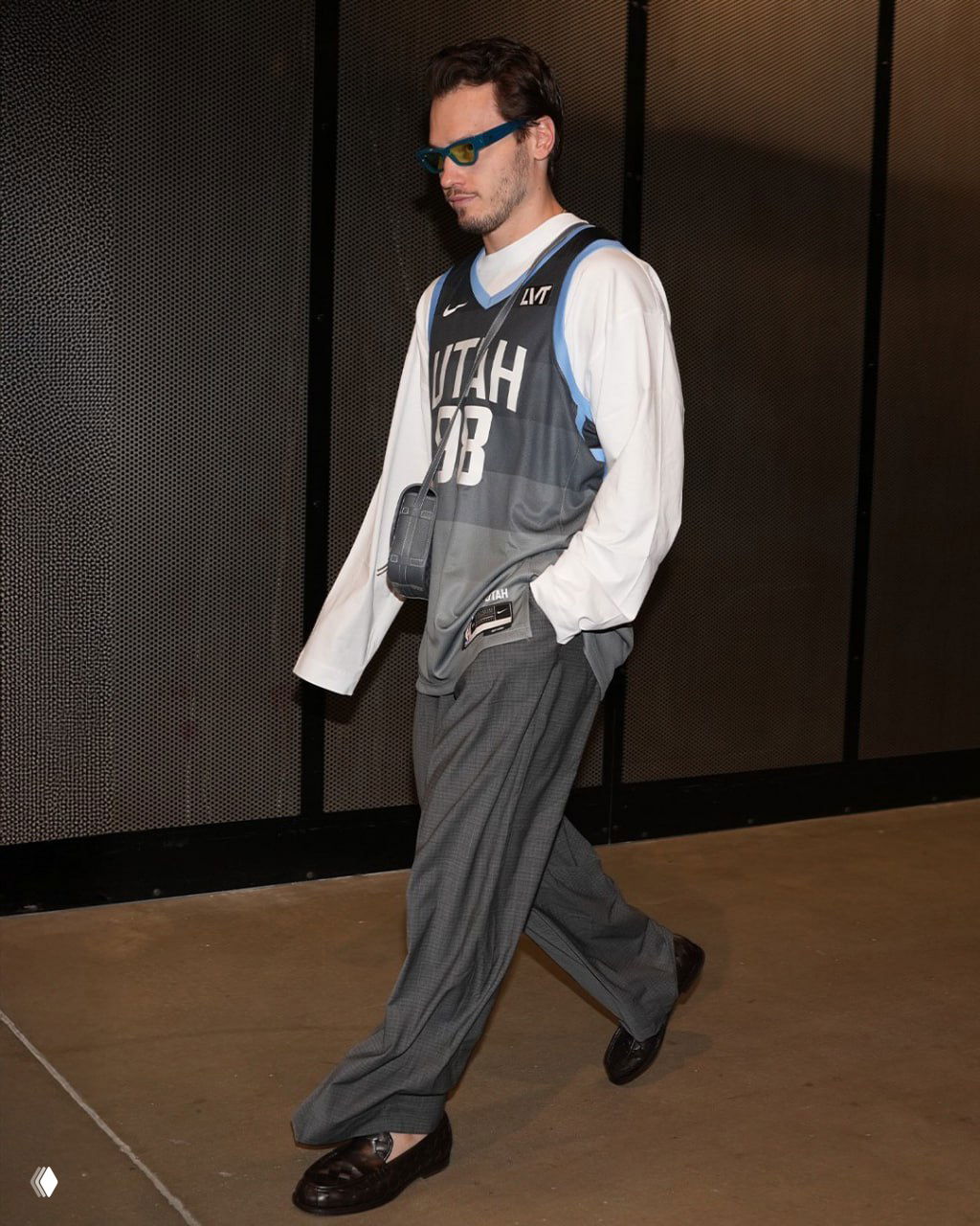 Человек в игровой майке «Utah» поверх длинного лонгслива и с небольшой сумкой через плечо идёт по коридору стадиона в цветных очках.