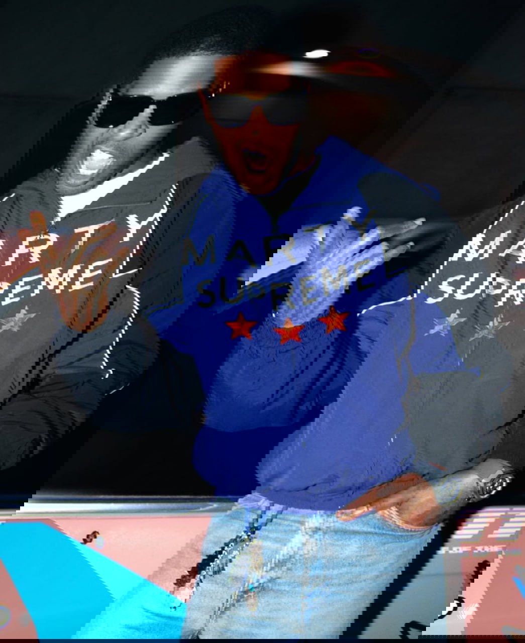 Ночной уличный кадр: человек в синей брендированной куртке демонстрирует жест рукой, видна надпись и звёзды на куртке.