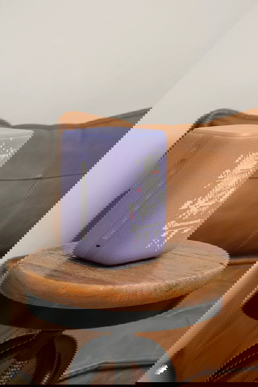 Фиолетовая Яндекс Станция 3 в лимитированном дизайне с абстрактной графикой, размещена на круглой деревянной табуретке рядом с креслом.