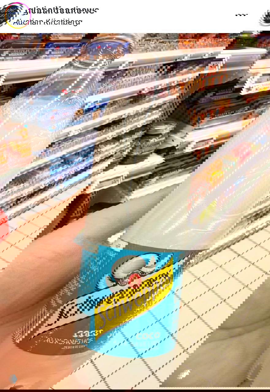 Крупный план бутылки Schweppes Coco с белой полупрозрачной жидкостью и изображением кокоса на этикетке на фоне прохода супермаркета.