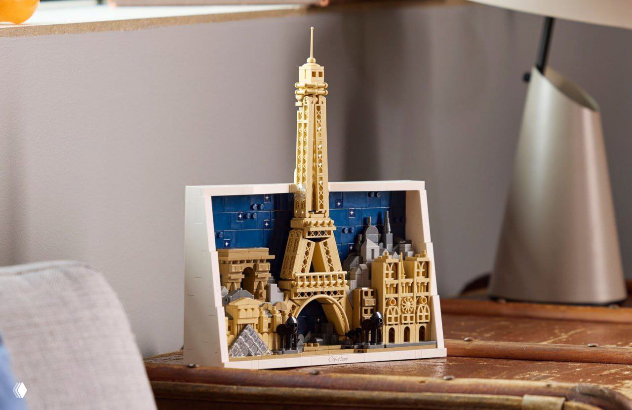 Собранный набор LEGO Paris установлен на деревянном столе рядом с лампой — видна вся композиция Эйфелевой башни, Лувра и Нотр‑Дама.