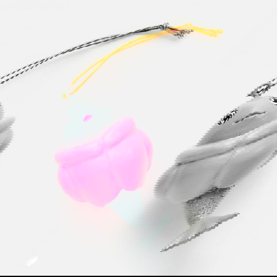 Маленькая ёлочная игрушка в форме рыбы в розовой «шубке», с серым хвостом и золотым шнурком, расположена на белом фоне.