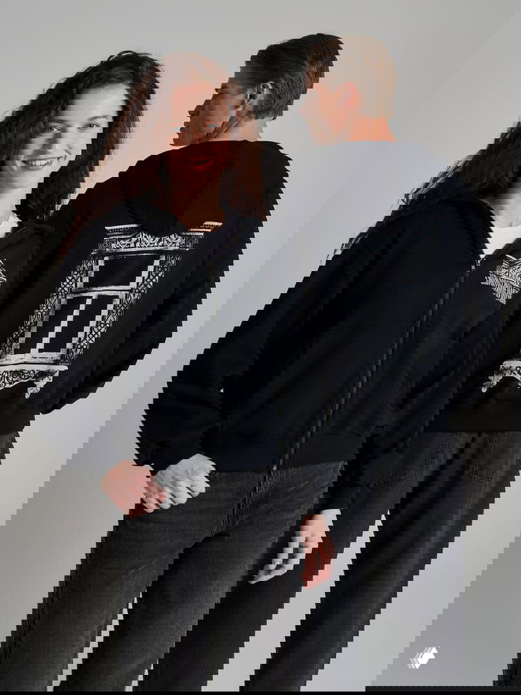 Модель в чёрной худи со стильным принтом деревянного наличника окна на спине и маленьким логотипом на груди.