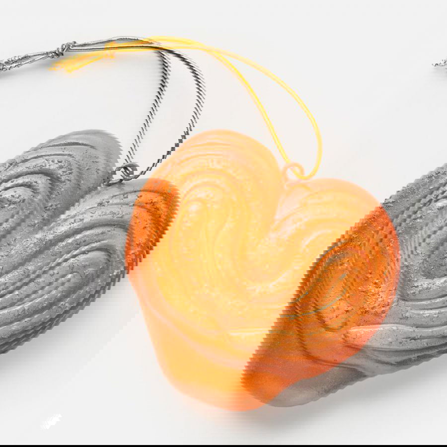 Оранжевая сердцевидная ёлочная игрушка в форме печенья или плюшки с рельефным узором и золотым шнурком, снята на нейтральном белом фоне.