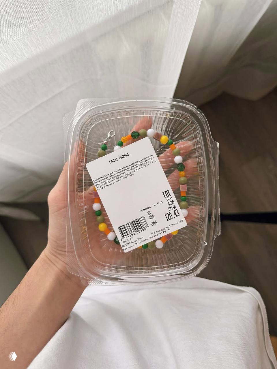Прозрачный пластиковый контейнер с наклейкой и внутри колье‑оливье из бусин; видно ценник и упаковку, как у салатной тары, съёмка сверху рукой.