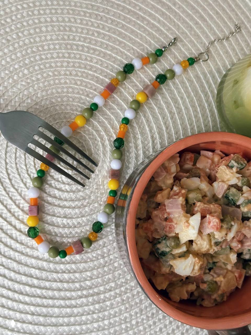 Колье-оливье рядом с миской салата оливье на текстурированной подставке; видны бусины, кусочки салата и части вилки в композиции.