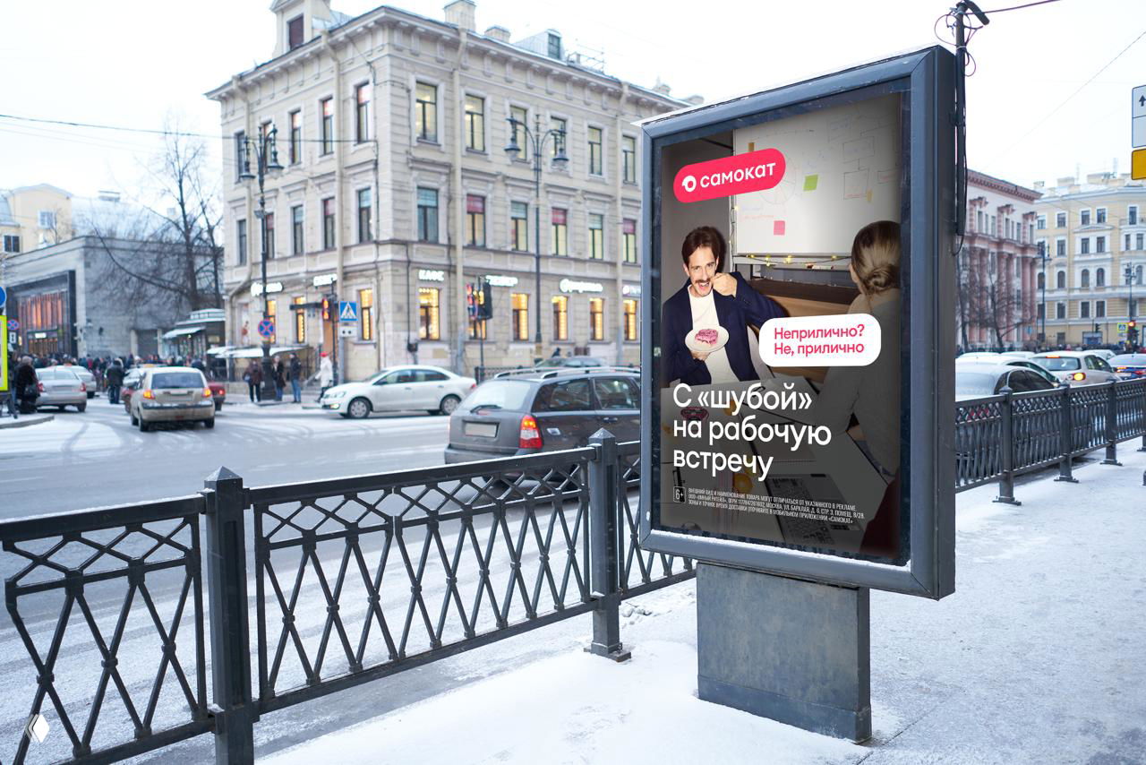 Городская стойка с постером Самоката зимой: уличная реклама на фоне городской улицы, на плакате человек с едой и слоган кампании.