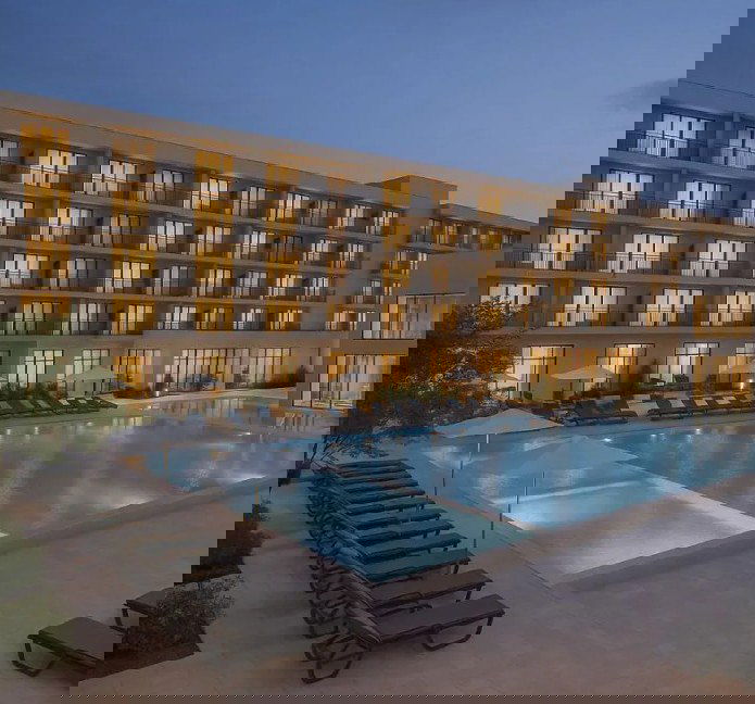 Ночной вид на современный отельный корпус с подсвеченным открытым бассейном и шезлонгами на переднем плане, зона отдыха у воды