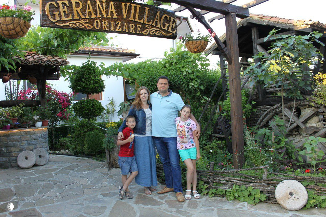Семья у вывески «GERANA VILLAGE ORIZARE»: родители с двумя детьми на фоне цветущего двора и декоративных элементов курортного комплекса.