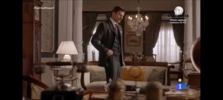 Кадр сцены: мужчина в костюме стоит в уютной гостиной с классической мебелью, люстрой и настольной лампой, интерьер домашнего пространства.