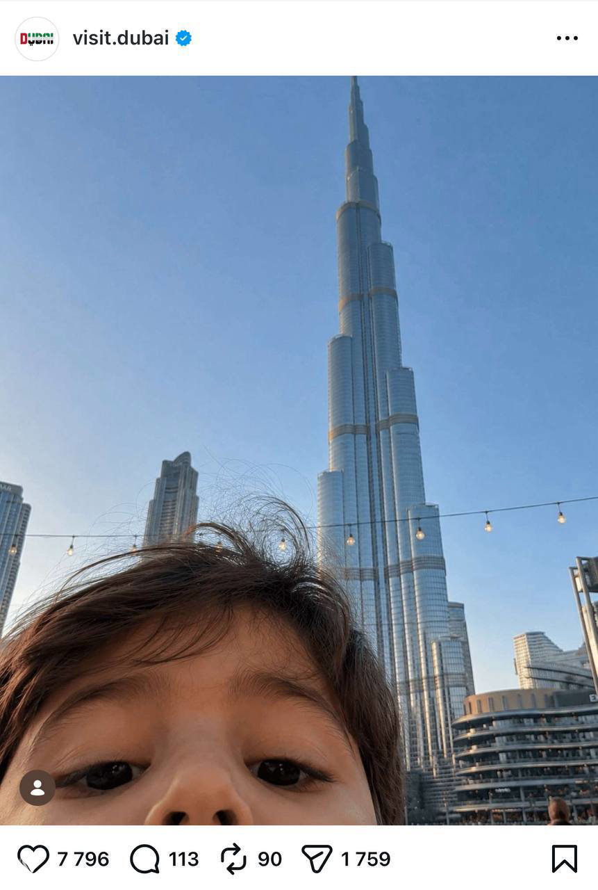 Детское селфи на фоне небоскрёба (Burj Khalifa) — ребёнок внизу кадра, городская панорама создаёт эффект случайной съёмки в публичном месте.