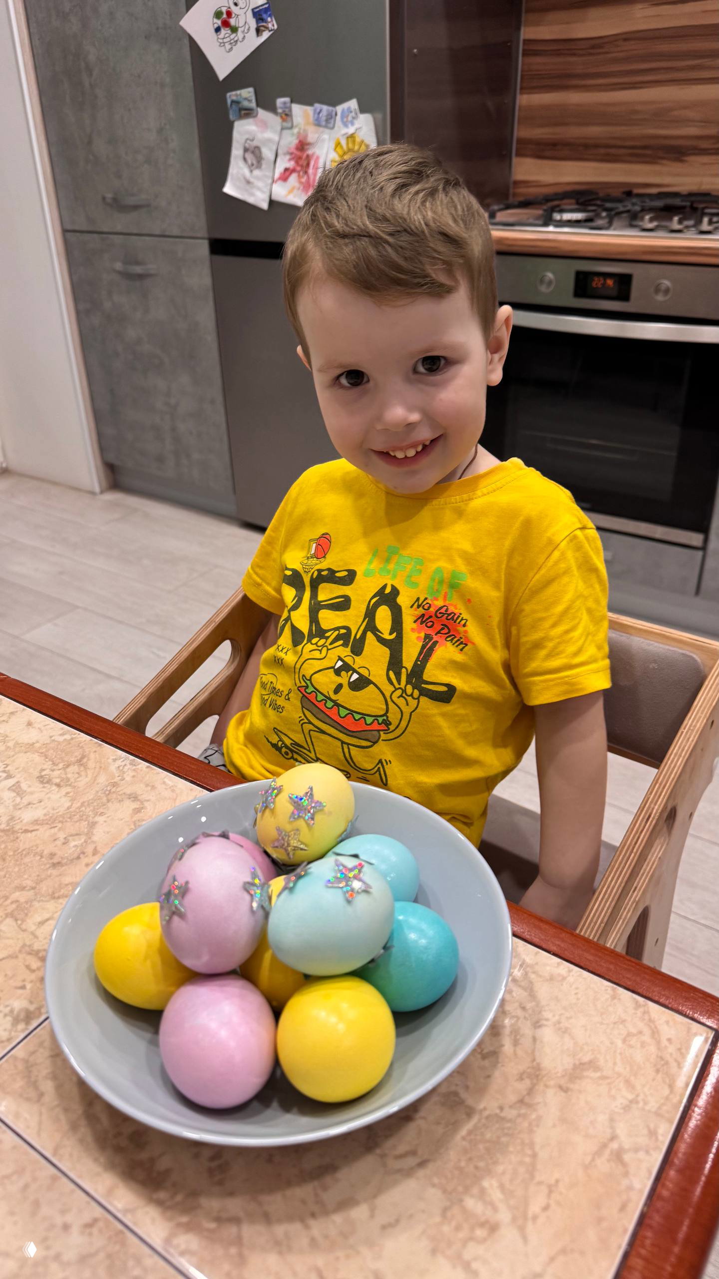 Улыбающийся ребёнок за кухонным столом рядом с миской пастельных окрашенных пасхальных яиц, украшенных блёстками и наклейками.
